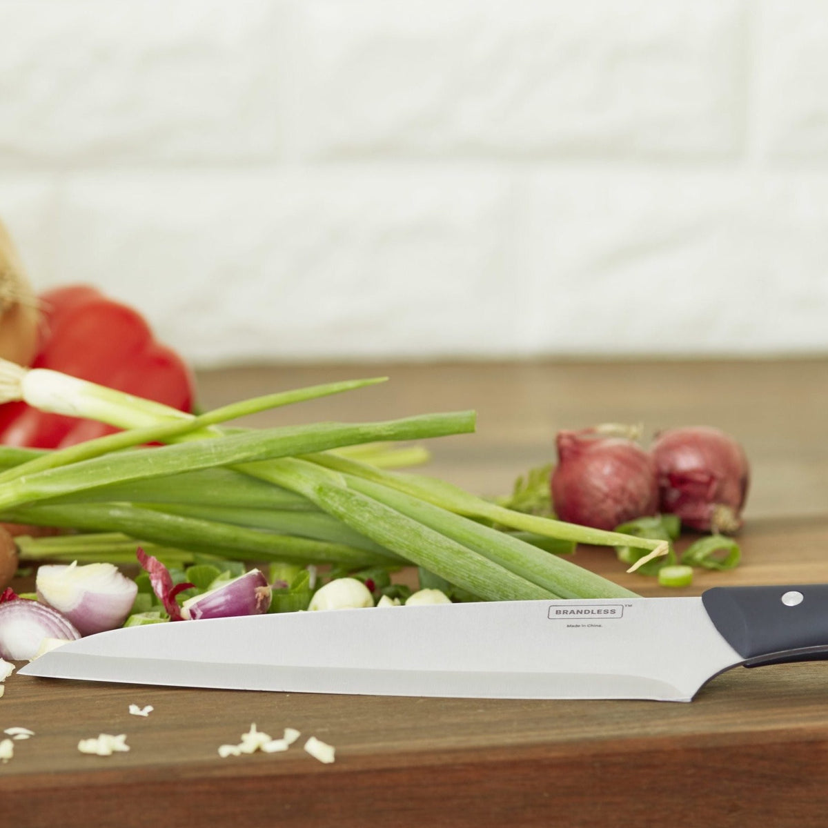 Lifestyle photo of the 8 inch chef&#39;s knife leaning on a cutting board with onions, shallots, and peppers on it.