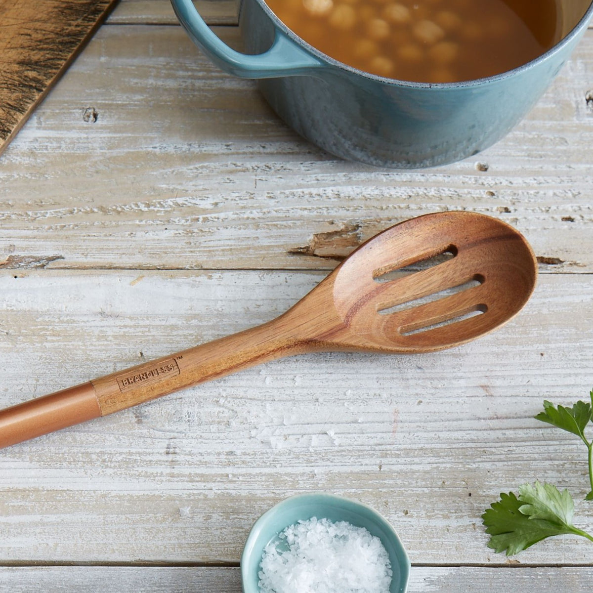 Lifestyle photo, acacia wood slotted serving spoon resting on kitchen table next to a pot full of chickpeas soaking.