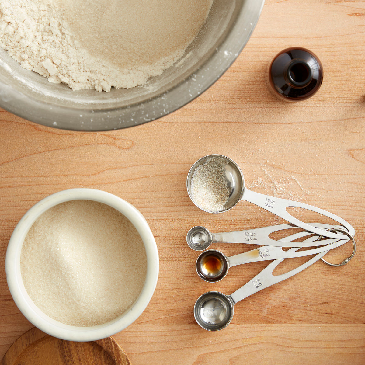 Lifestyle photo, showing a baker creating cookie dough, with the different sizes of measuring spoon showing the leftovers of flower, sugar, vanilla extract, and salt.