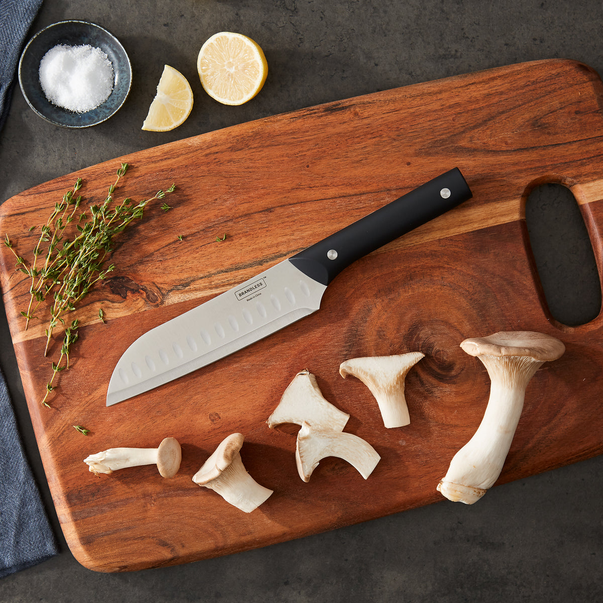 Lifestyle photo, santoku knife laying on wood cutting board with large-sliced mushrooms, wedged lemons, coarse salt, and fresh seasonings.