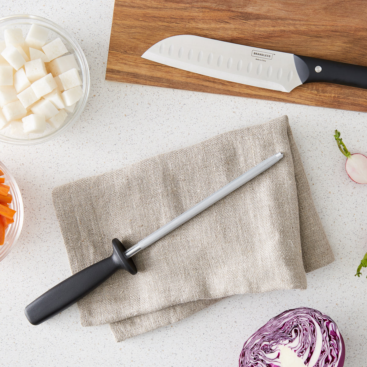 Lifestyle photo, honing steel laying on a dishcloth on a counter next to diced and sliced veggies and a santoku knife.