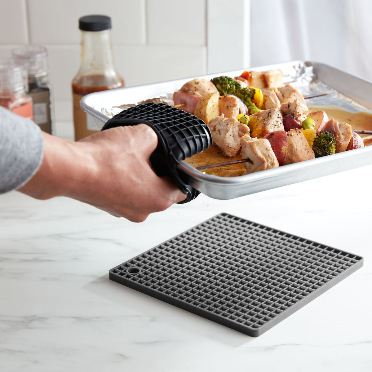 Lifestyle. Hand placing a fresh-off-the-grill pan full of chicken skewers down onto a gray trivet protecting a marble countertop