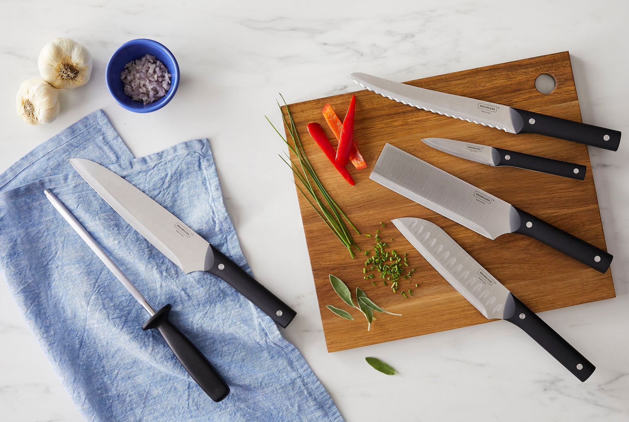 Lifestyle image of an array of kitchen knives laying on a cutting board next to some spices, sprigs, and peppers.