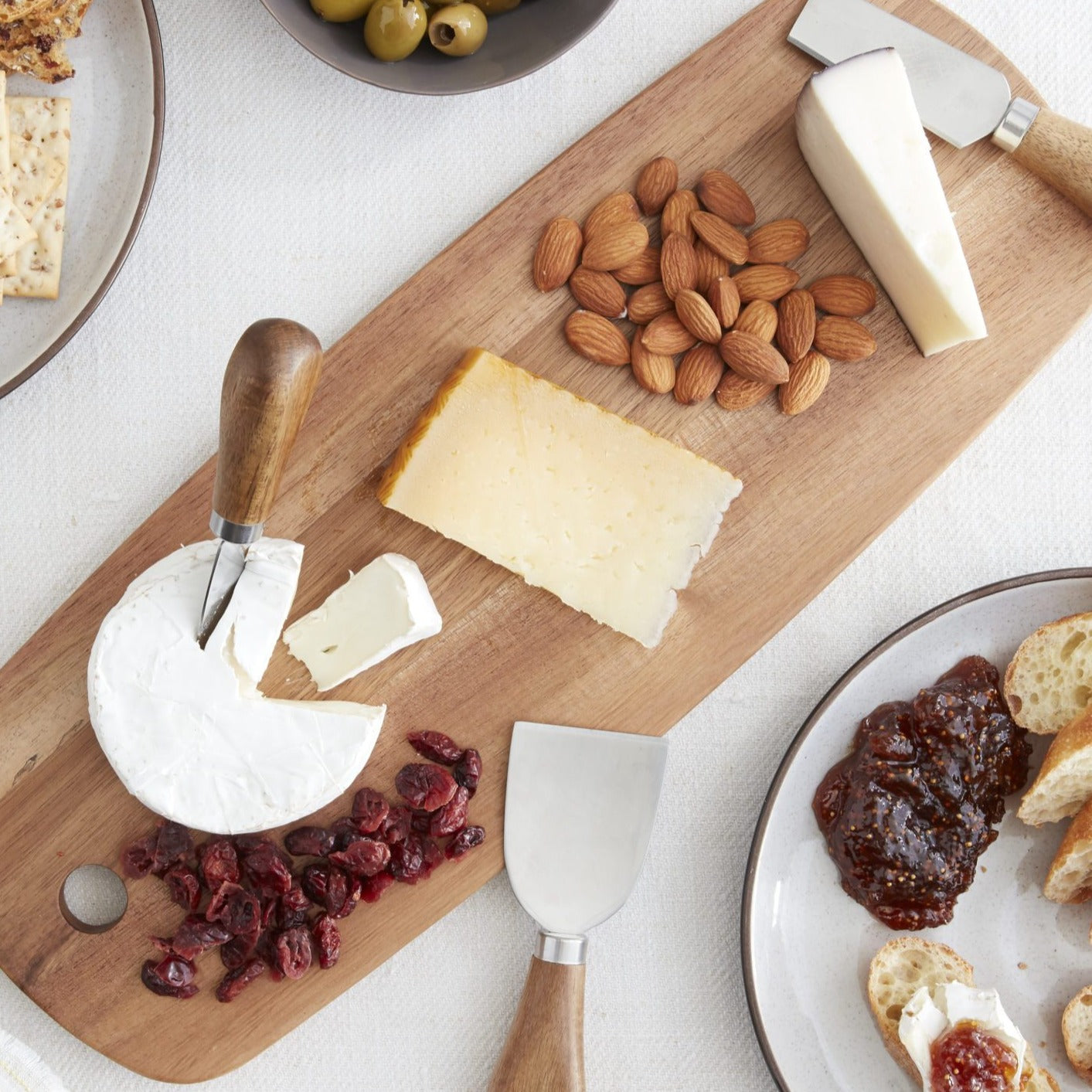 Lifestyle photo, acacia cheese board with tapas preparation ingredients including dried cranberries, three types of cheese, and almonds.