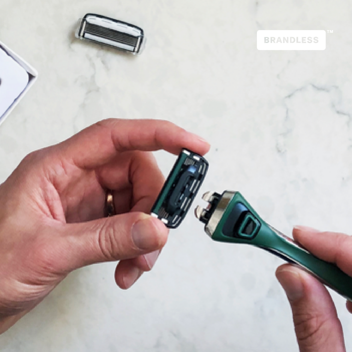 Lifestyle photo, man assembling the razor by clicking the weighted handle into the back of the razor cartridge for face, background is a white marble bathroom countertop, brandless logo.