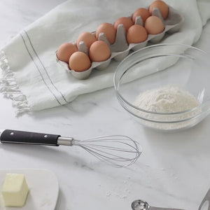 Video, no sound. Woman mixes flour and melted butter in a glass mixing bowl next to a carton of eggs, a pad of butter, and a set of brandless measuring spoons.