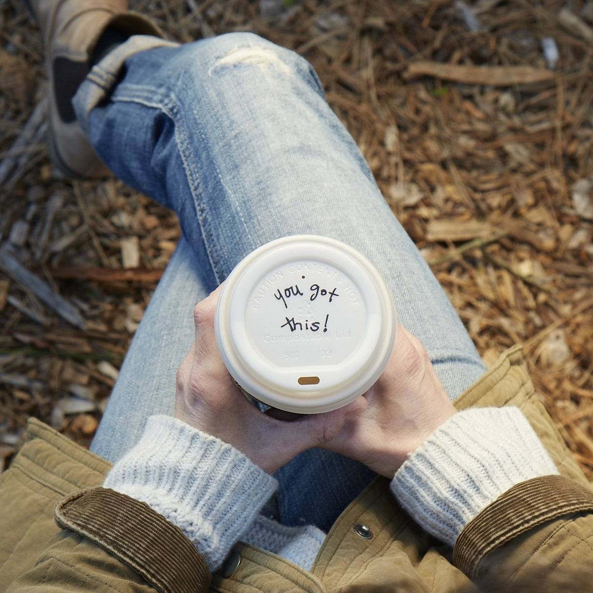 Lifestyle photo of a person holding a cup of coffee while seated in a park with the inspirational message of  &#39;you got this!&#39; written ono the lid.