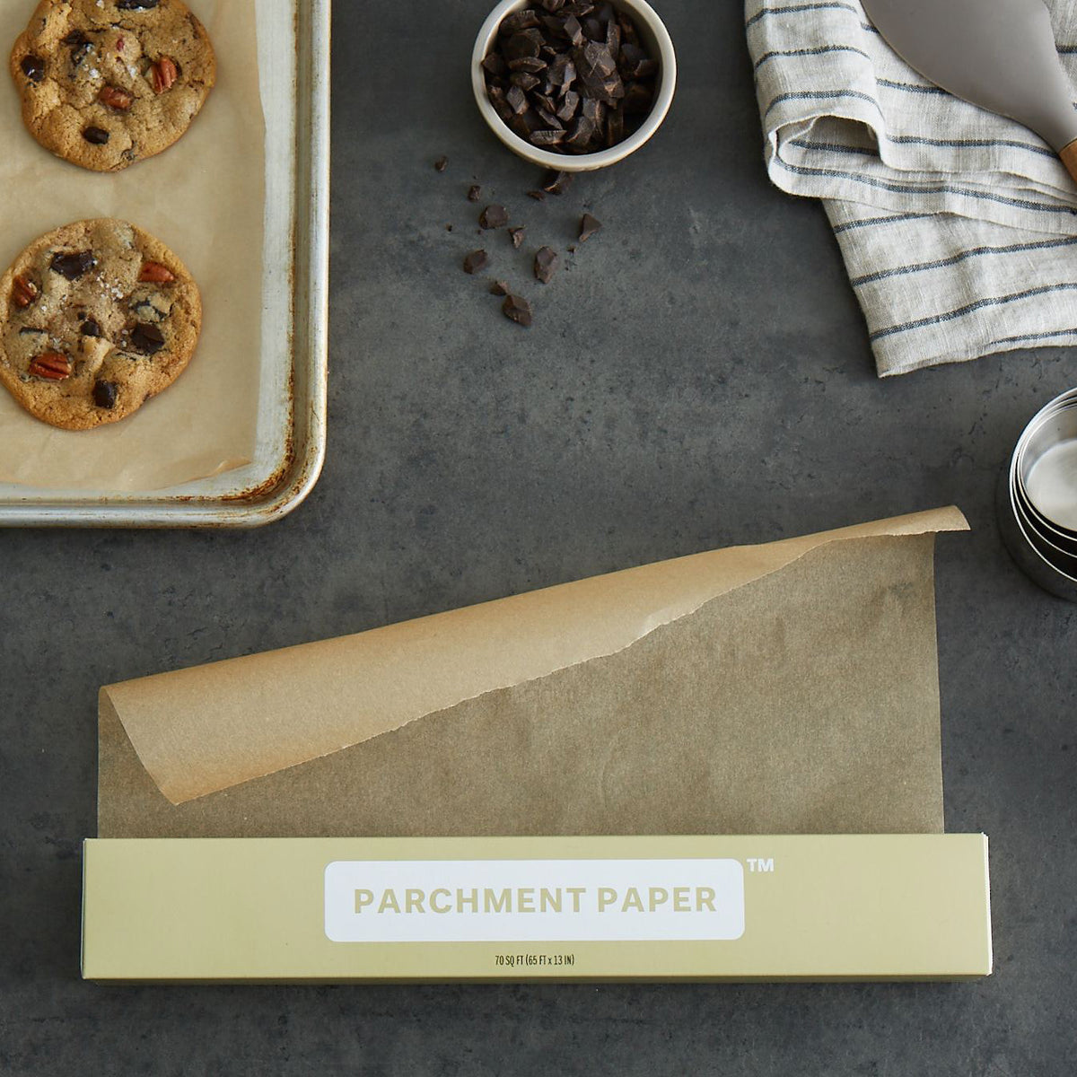 Lifestyle photo, parchment paper unfurled from box next to a baking tray lined with parchment paper with freshly baked chocolate chip and pecan cookies.