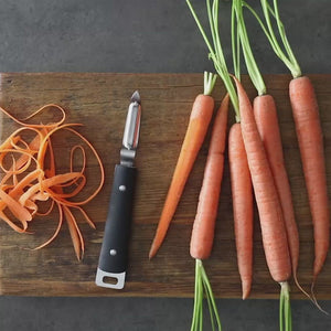 Video, no sound. Woman uses brandless vegetable peeler to effortlessly shave uniform strips of carrot off a fresh carrot from the garden onto a cutting board.