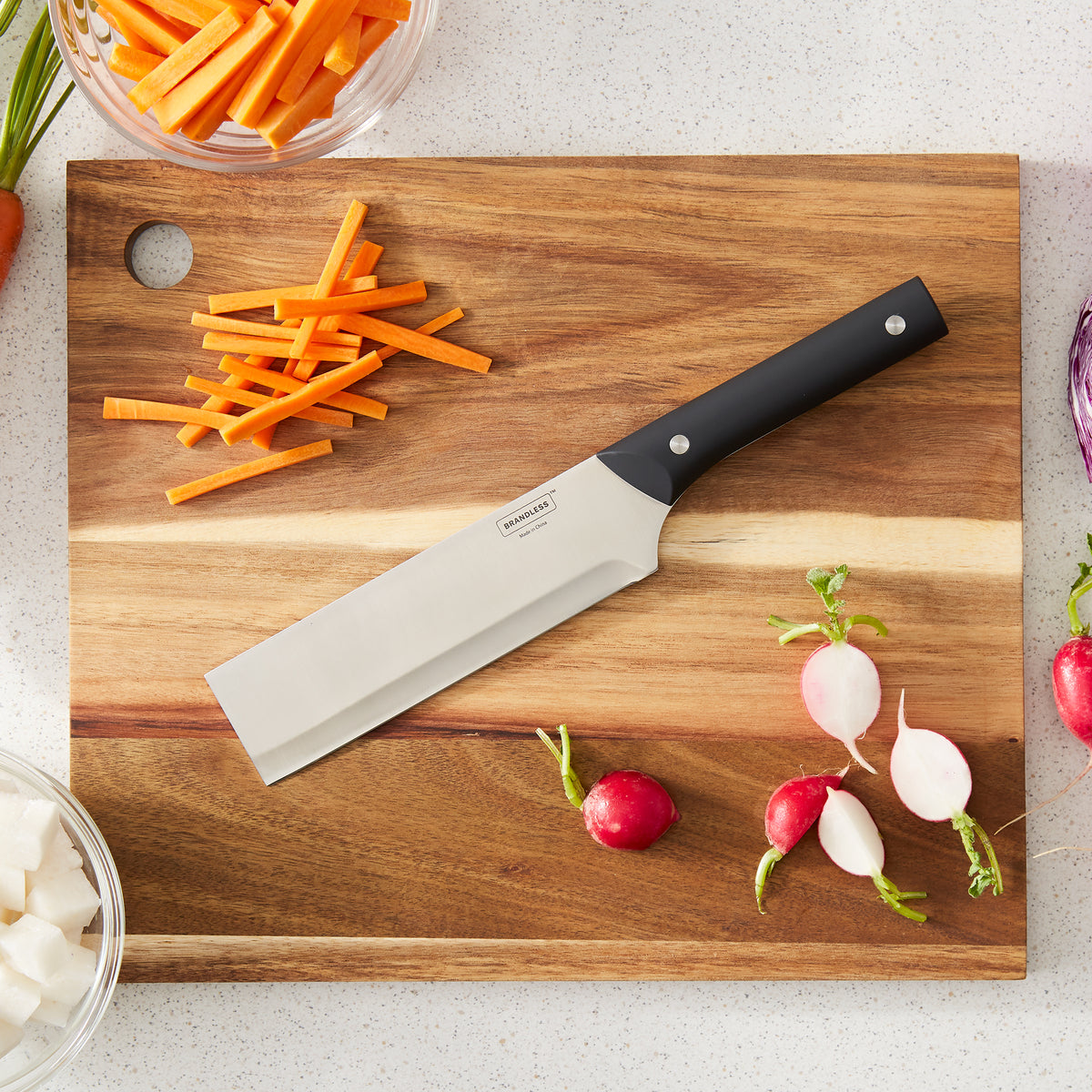 Lifestyle photo, showing nakiri knife laying on a wood cutting board with chopped carrots and quartered radishes.