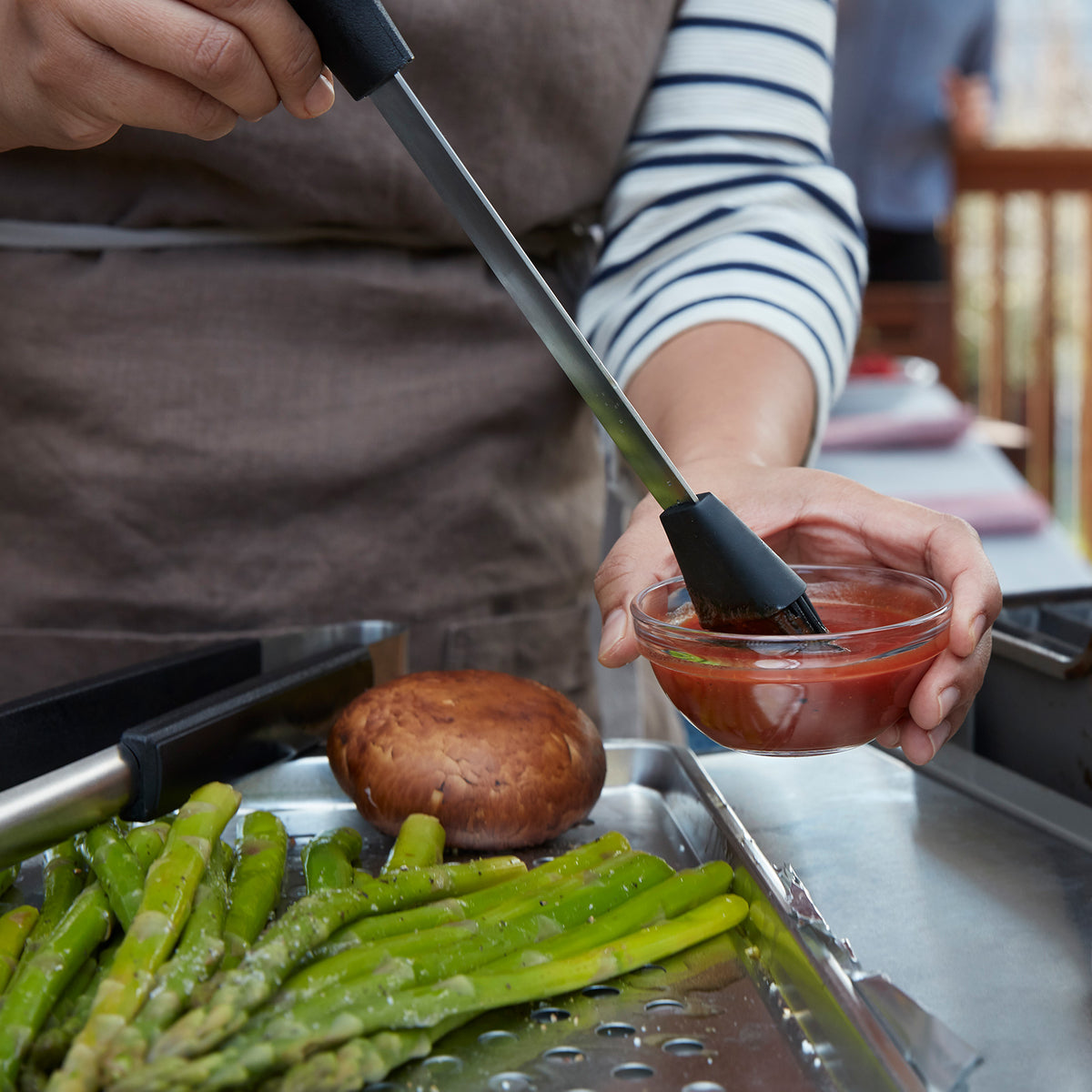 Lifestyle photo, a man preparing to add barbeque sauce to some items on the grill, seen next to fresh asparagus.