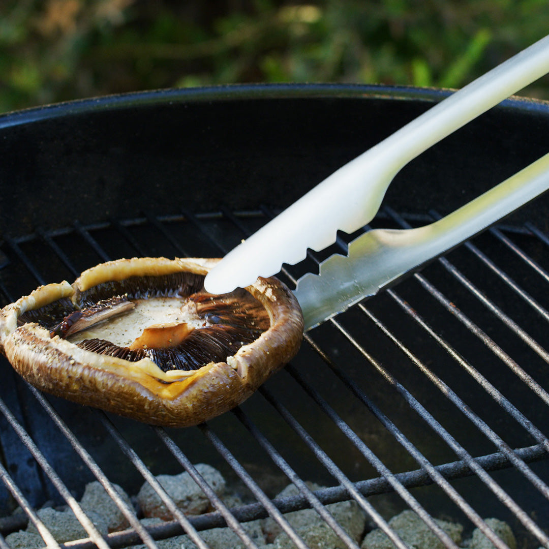 Lifestyle photo, close up shot of the scalloped tips of the grill tongs picking up a portabello mushroom cap off a charcoal grill.