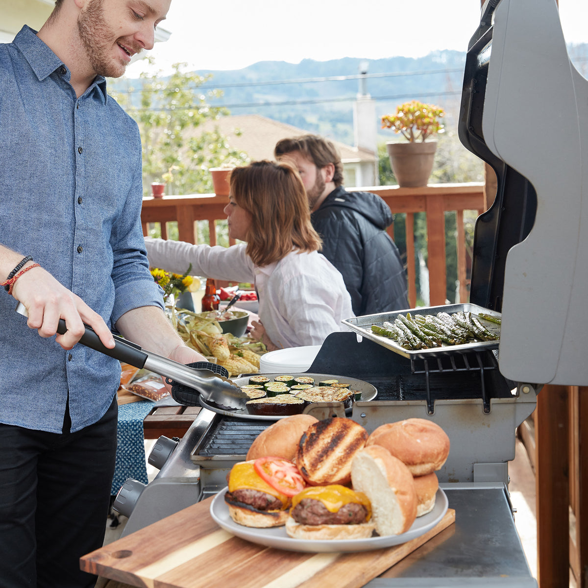 Lifestyle photo, a man using the grill tongs to transfer roasted vegetable slices from the grill to a serving platter, while guests in the background enjoy the barbeque.