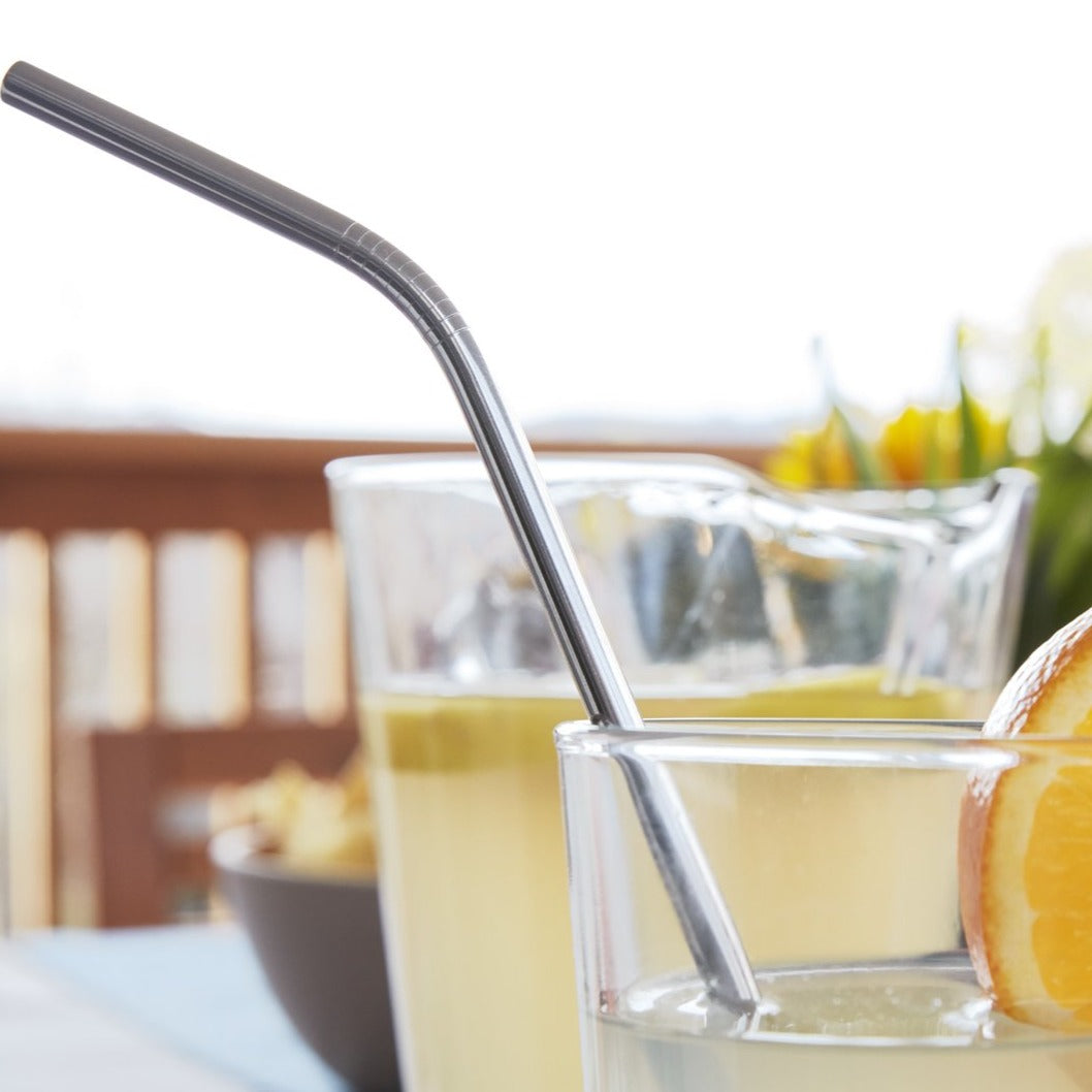 Lifestyle photo, a summertime lemonade in a tall glass tumbler with a fresh orange slice using a brandless stainless steel straw sits on an outdoor patio table.