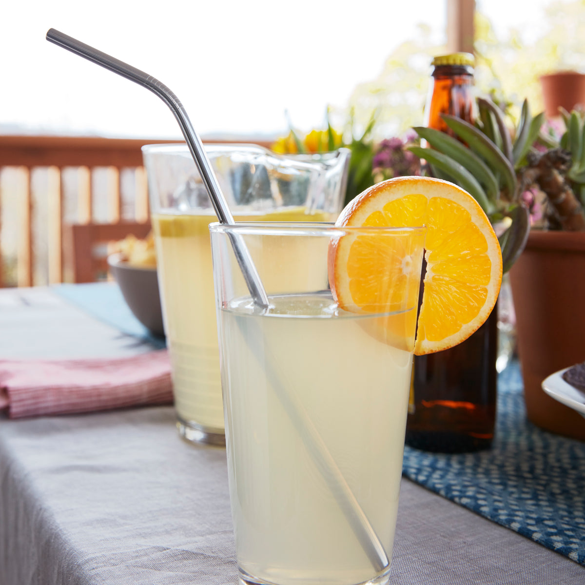 Lifestyle photo, a summertime lemonade in a tall glass tumbler with a fresh orange slice using a brandless stainless steel straw sits on an outdoor patio table.