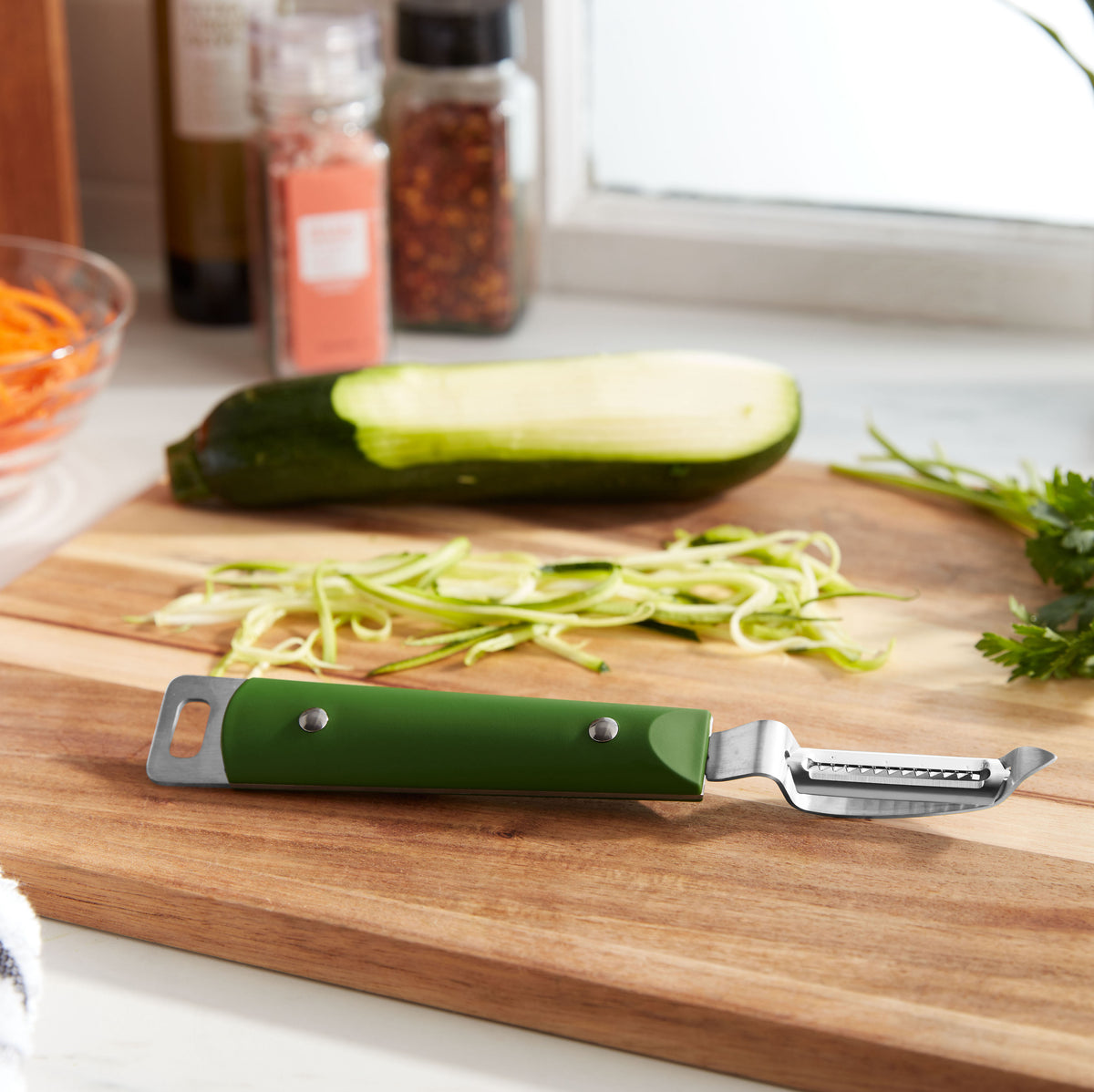 Lifestyle photo showing green-handled julienne peeler on a cutting board next to a zuchinni with fresh zoodles having been effortlessly just created.