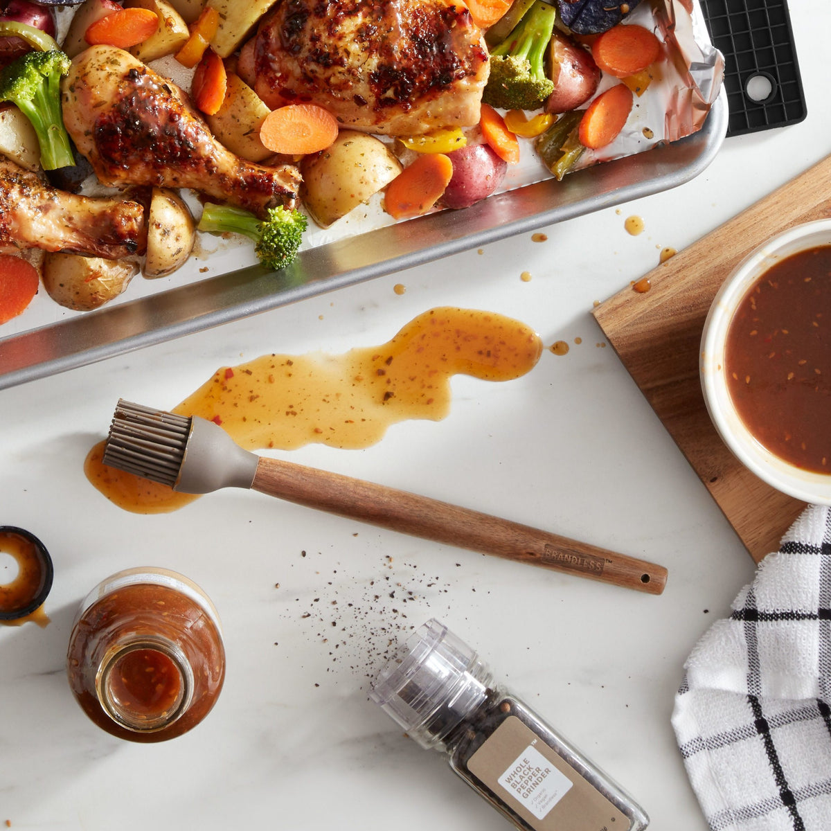 Lifestyle photo, silicone basting brush on a kitchen counter having just spread a sweet chili marinade over a baking tray of chicken separates and chopped veggies.