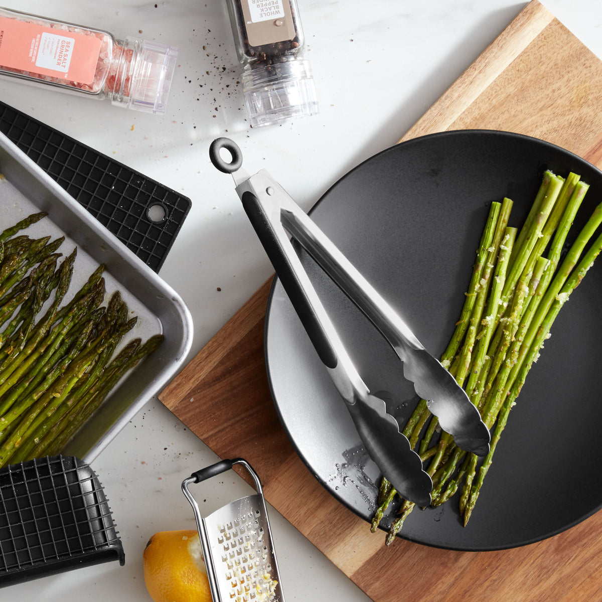Lifestyle photo, 9 inch stainless steel tongs locked in a semi-closed position, leaning on the edge of a plate having just transferred some roasted asparagus spears from a baking tray to a dinner plate.