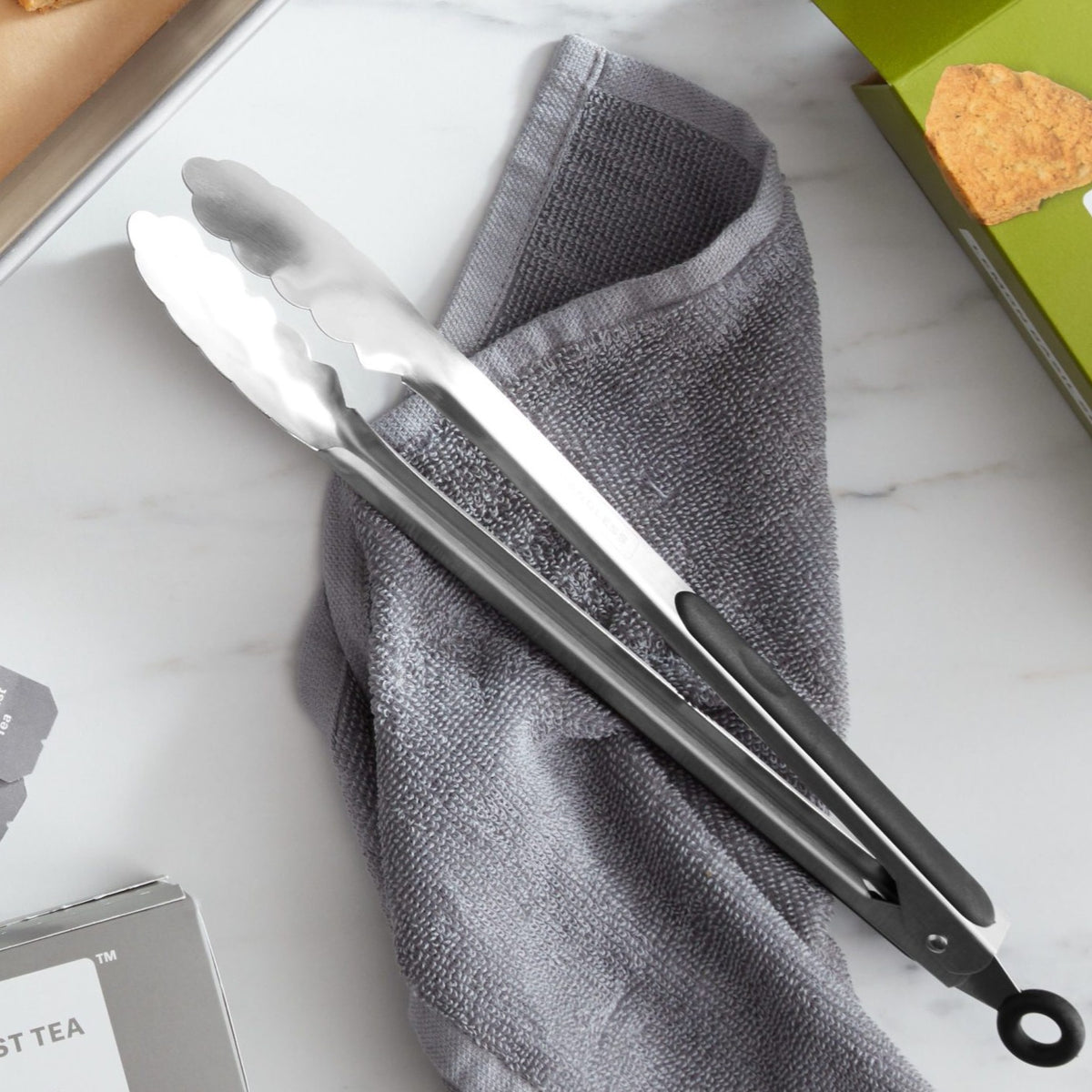 Lifestyle photo, side view, 12 inch stainless steel tongs, in closed and locked position, laying on kitchen towel on a kitchen counter.