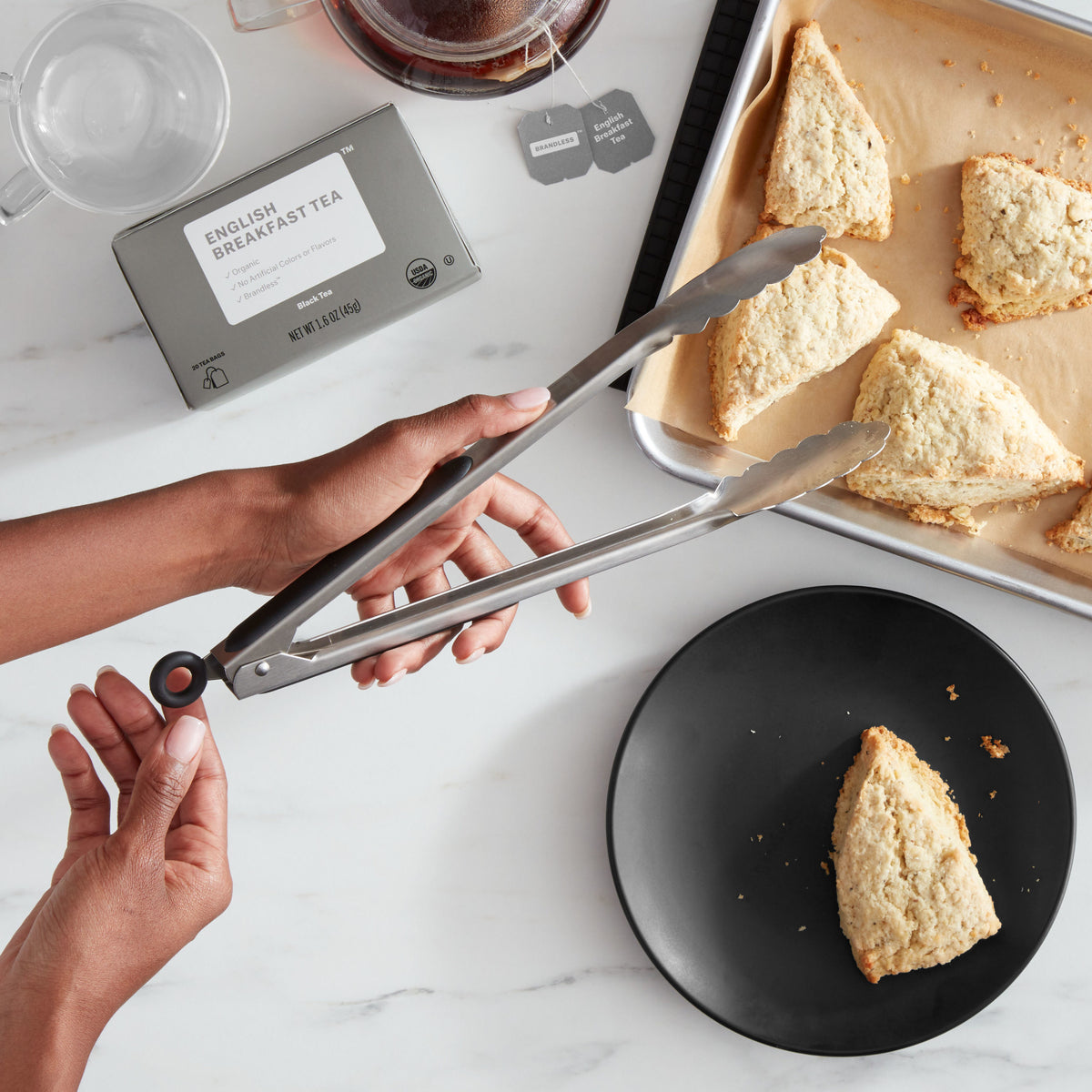 Lifestyle photo, woman&#39;s hands holding 12 inch stainless steel tongs and transferring fresh scones from a baking tray to a plate on a marble kitchen countertop.