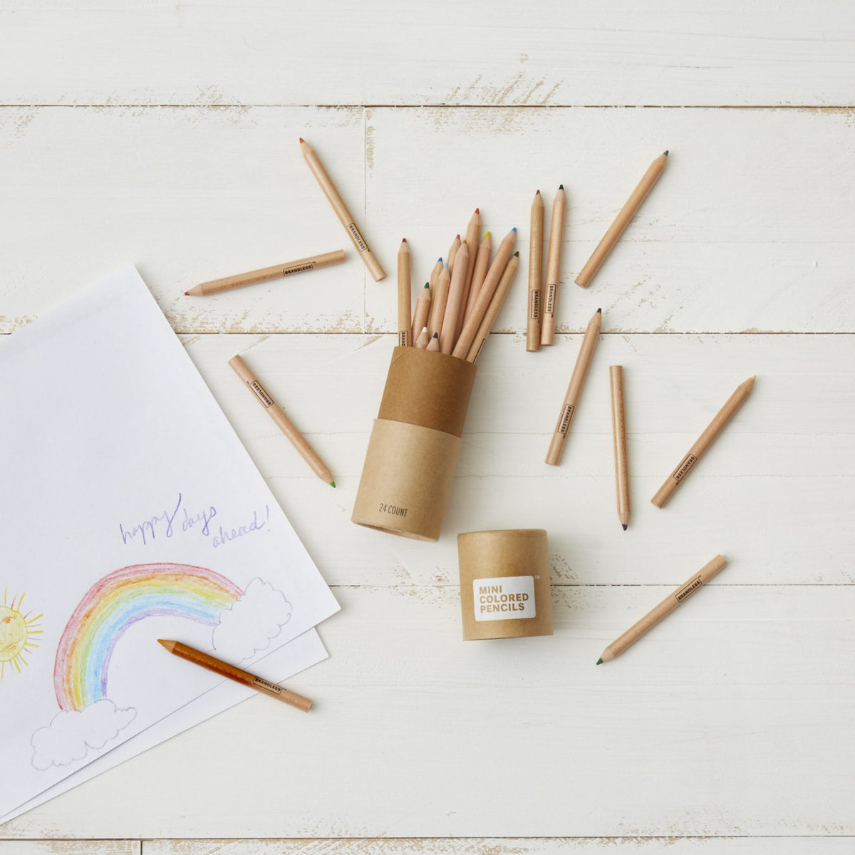 Lifestyle photo, showing a child&#39;s drawing of a rainbow next to a canister of mini colored pencils with many colors strewn about during the creative process of drawing.