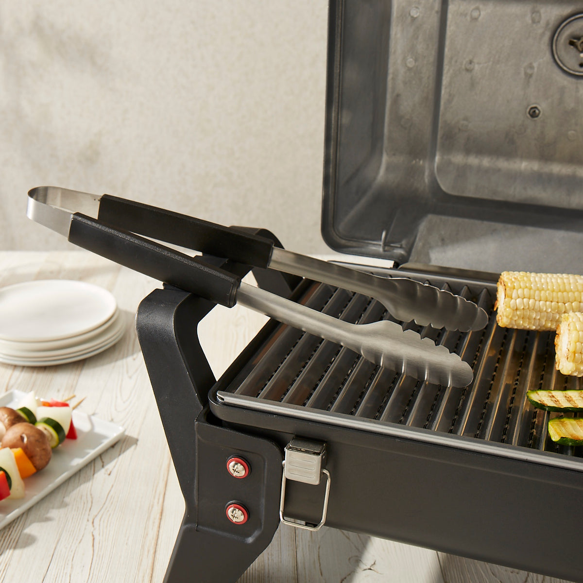 Lifestyle photo, grill tongs resting on the side of a countertop mini grill ready to serve up some roasted corn on the cob and zucchini fries.