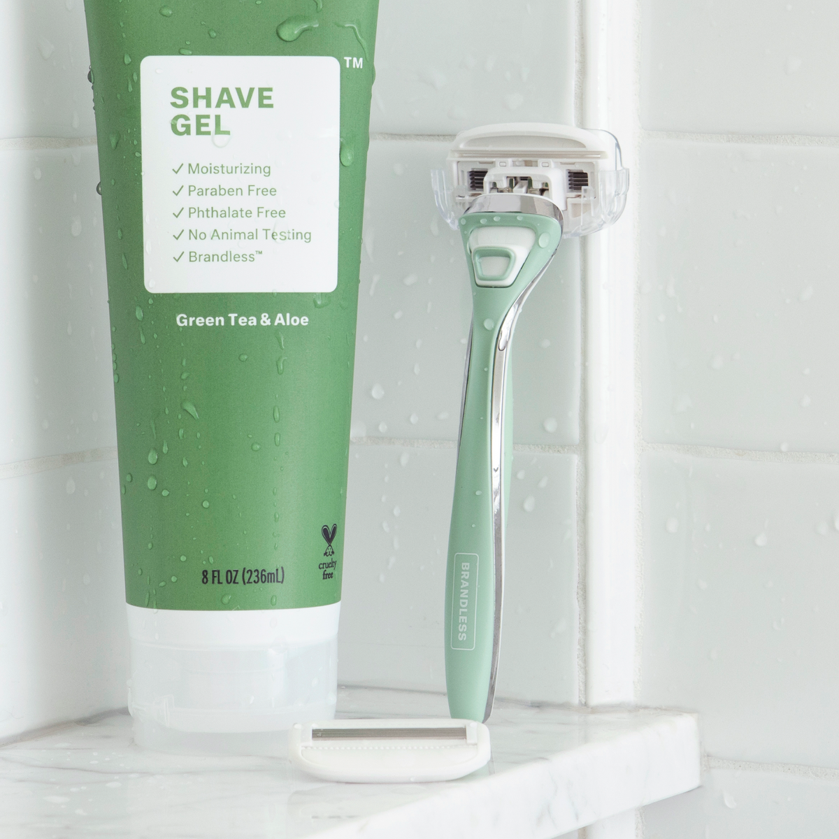 Lifestyle photo, razor for body stands up against the tile wall of a shower on a shelf next to a tube of brandless green tea and aloe shave gel.  Water droplets abound.