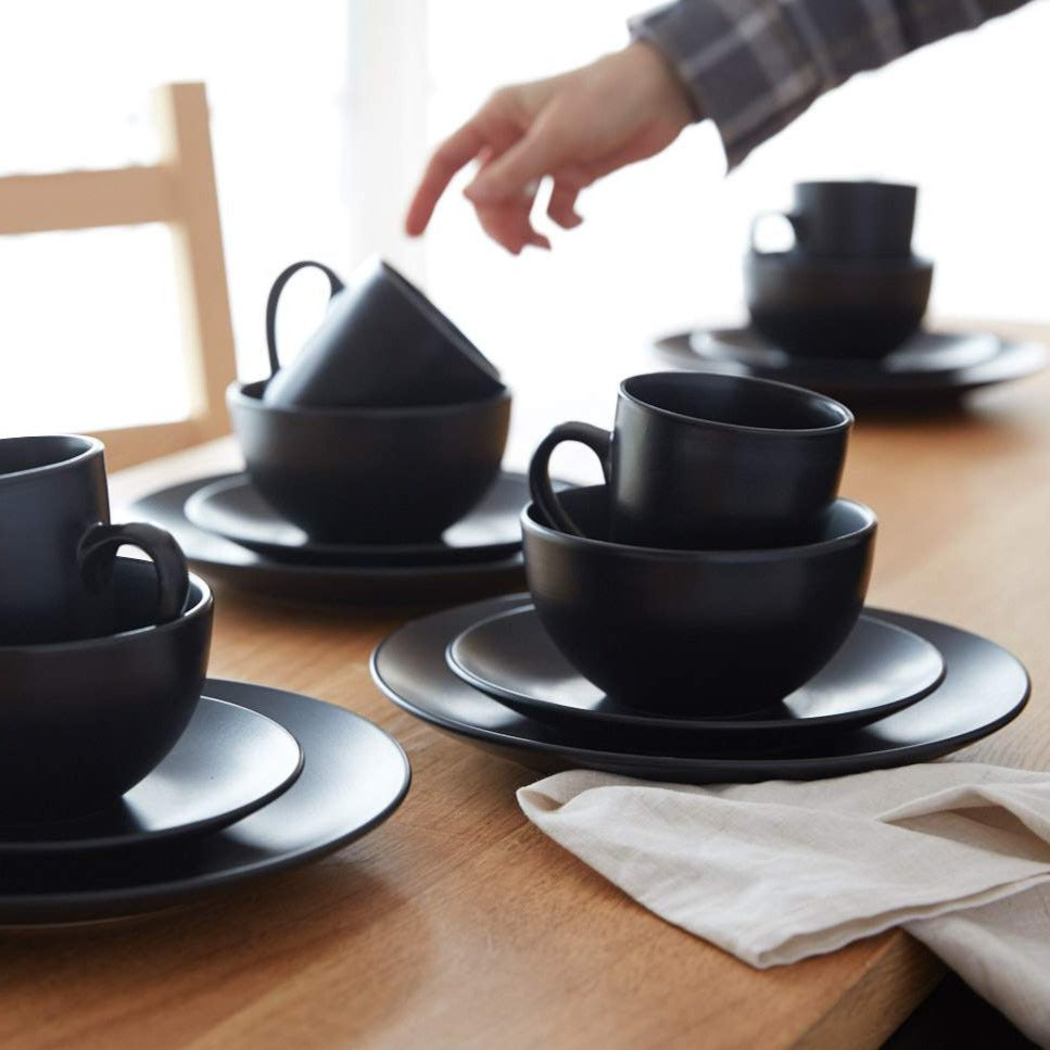 Lifestyle photo, stacked stoneware service sets of a dinner plate, salad plate, cereal bowl, and mug. Three piles of stoneware decorate a kitchen table and a hand reaches in to pick up a mug.